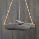 The Earthy Anthracite Bamboo Hanging Bird Bath/Feeder W23.3cm is a gray, frost-proof ceramic feeder with a bird figurine in the center, suspended by ropes against dark wood.