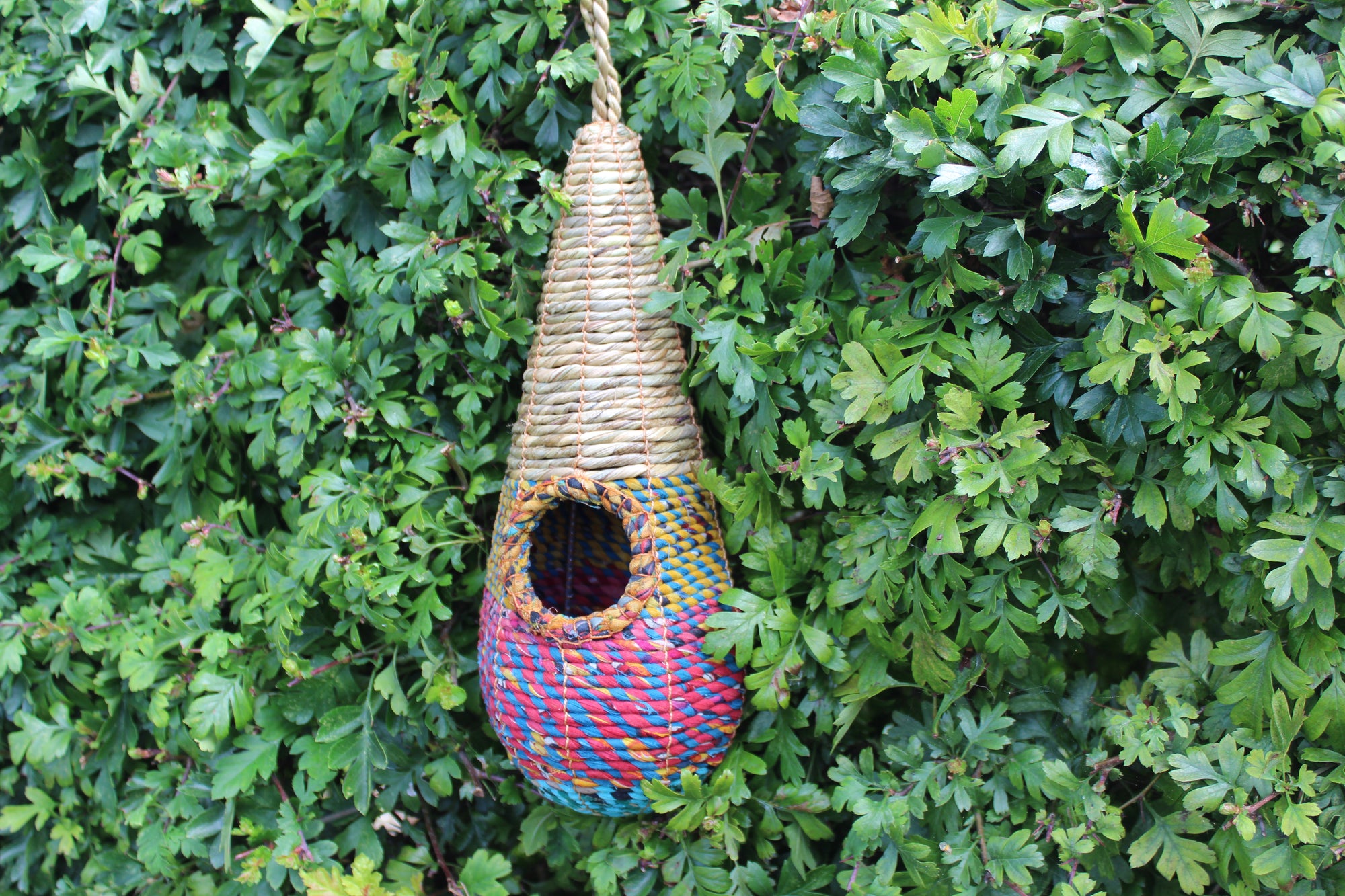 A vibrant multicolored woven birdhouse, shaped like a tear drop, hangs from a rope against a lush green leafy background. The Wildlife World Tahera Artisan Wild Bird Nester—an elegant Fair Trade design with its small circular entrance near the top—showcases artisan handcrafted quality in its eye-catching fibers.