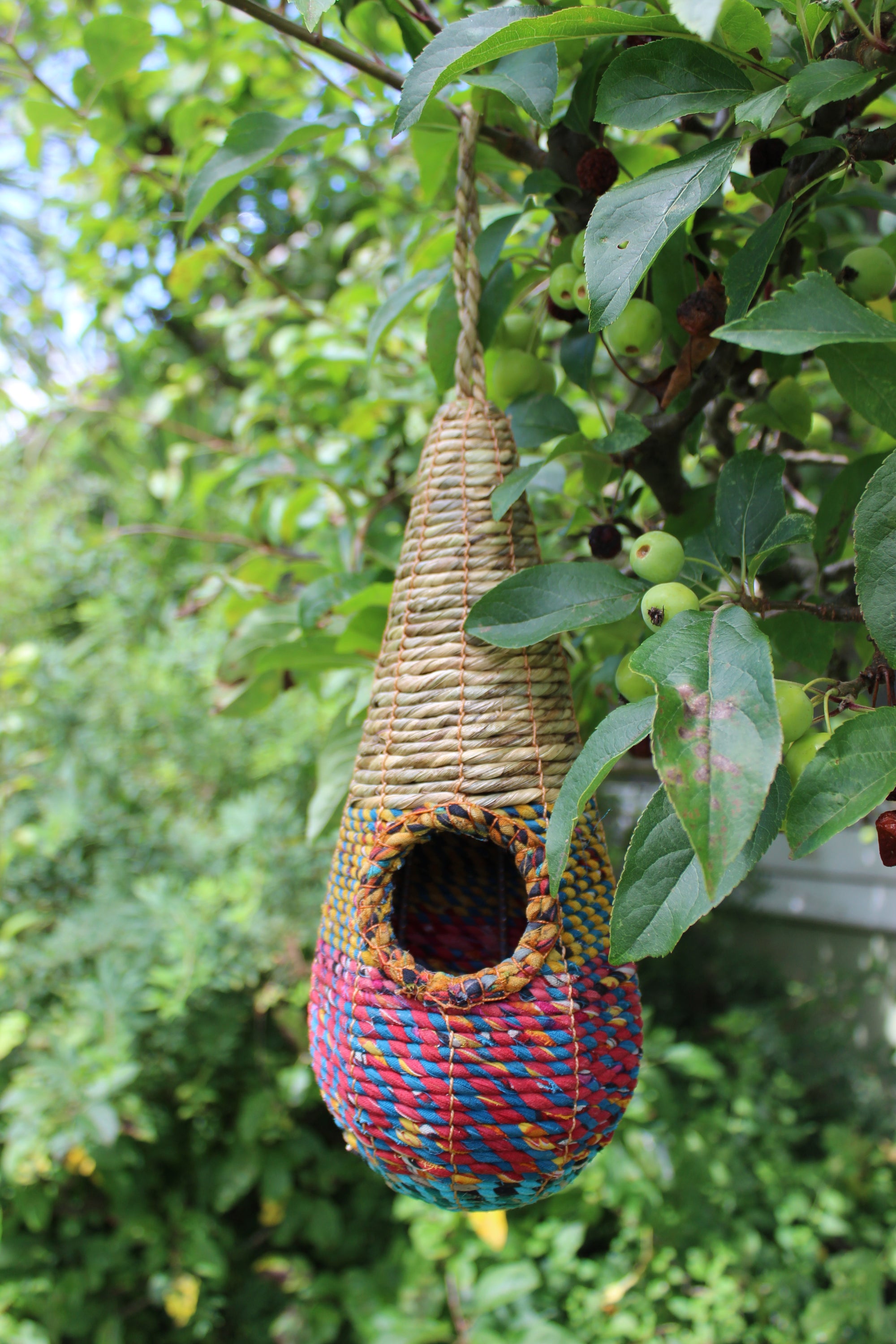 The Wildlife World Tahera Artisan Wild Bird Nester - Elegant Fair Trade Teardrop Design, with its intricate natural, red, blue, and yellow weaving patterns, hangs gracefully from a leafy tree branch. The teardrop-shaped birdhouse features a round entrance and is set against a backdrop of green foliage and small unripe apples.