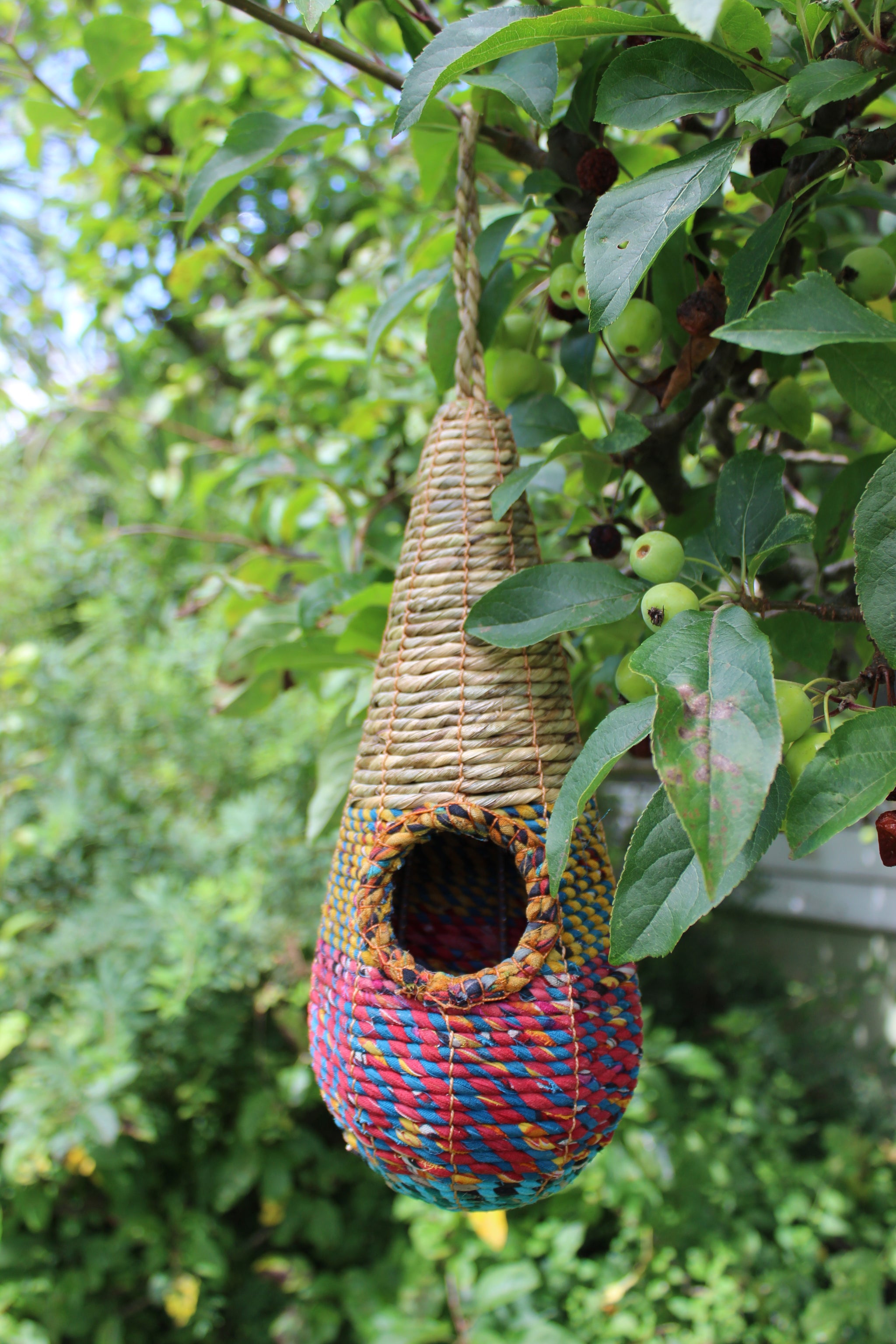 The Wildlife World Tahera Artisan Wild Bird Nester - Elegant Fair Trade Teardrop Design, with its intricate natural, red, blue, and yellow weaving patterns, hangs gracefully from a leafy tree branch. The teardrop-shaped birdhouse features a round entrance and is set against a backdrop of green foliage and small unripe apples.