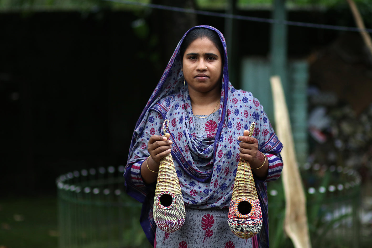 A woman adorned in a patterned blue and purple scarf and dress stands outdoors, holding two intricately woven, colorful bird nests. Demonstrating her expertise as the Tahera Artisan behind the Wildlife World Fair Trade Teardrop Wild Bird Nester, she poses against a backdrop of blurred greenery and a fenced area.