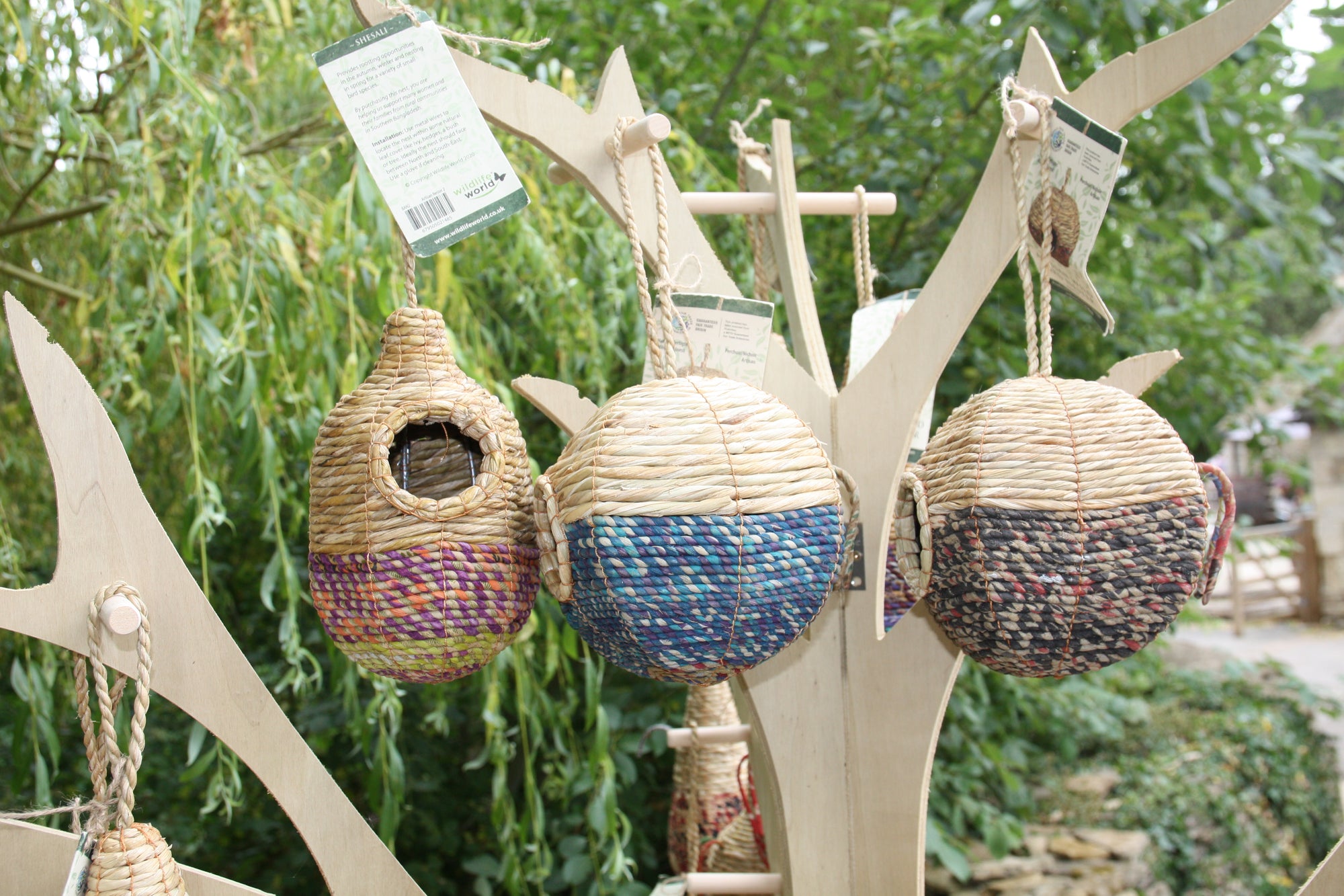 Three Wildlife World Tahera Artisan Wild Bird Nester houses, featuring elegant fair-trade teardrop designs, hang from the branches of a simple wooden stand outdoors. Each handcrafted bird house varies in design, with two showcasing colorful patterns. Trees and greenery in the background highlight their artisan quality.