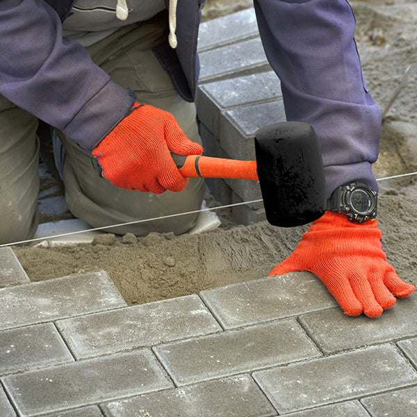 A person wearing red gloves uses a rubber mallet to adjust concrete pavers. Dressed in a blue jacket and beige pants, they kneel on the ground while laying the pavers. A Dekton 225Ft Masonry Brick Line 12pc CDU guides their alignment efforts, reminiscent of those seen in construction projects.