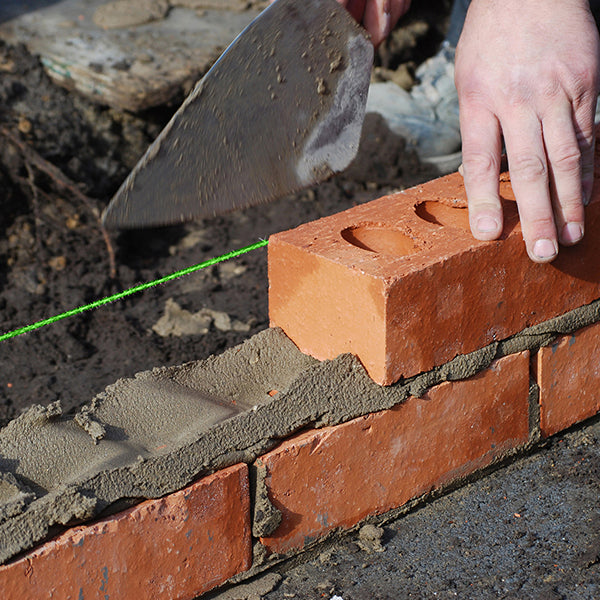 A close-up of a person laying a brick in a wall under construction. The individual uses a trowel to apply mortar to the top of the previous layer of red bricks, aligning the new brick with a Dekton 225Ft Masonry Brick Line from the 12pc CDU. The mortar is grey, commonly found in construction projects.