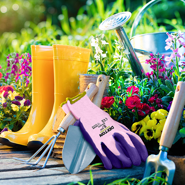A vibrant garden scene featuring yellow rain boots, a watering can, and various gardening tools, including Dekton Tough Grip Latex Coated Gardening Gloves 7/Small, a trowel, and a cultivator set amongst blooming flowers and greenery. The overall setup rests on wooden decking, capturing a sunny outdoor setting.