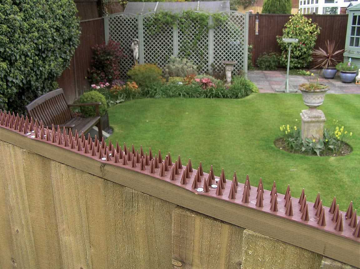 A garden surrounded by a sturdy fence adorned with Fence Guard Plastic Garden Security Spikes (1 x Strip, Brown) showcases green grass, vibrant flower beds, and a welcoming bench. Lattice panels, a bird feeder, and charming potted plants sit in the background.