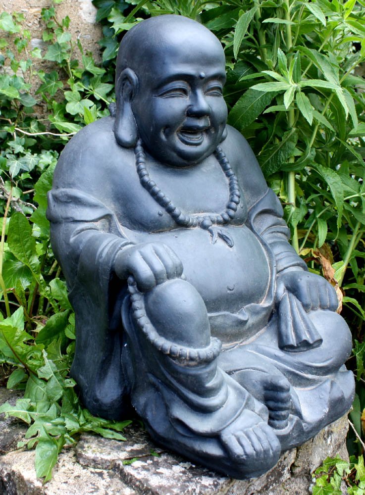 The Stone Effect Laughing Buddha Statue, with its black finish and joyful expression, sits with one knee raised in the zen room. The statue features a round belly and holds prayer beads in one hand, surrounded by lush green plants.
