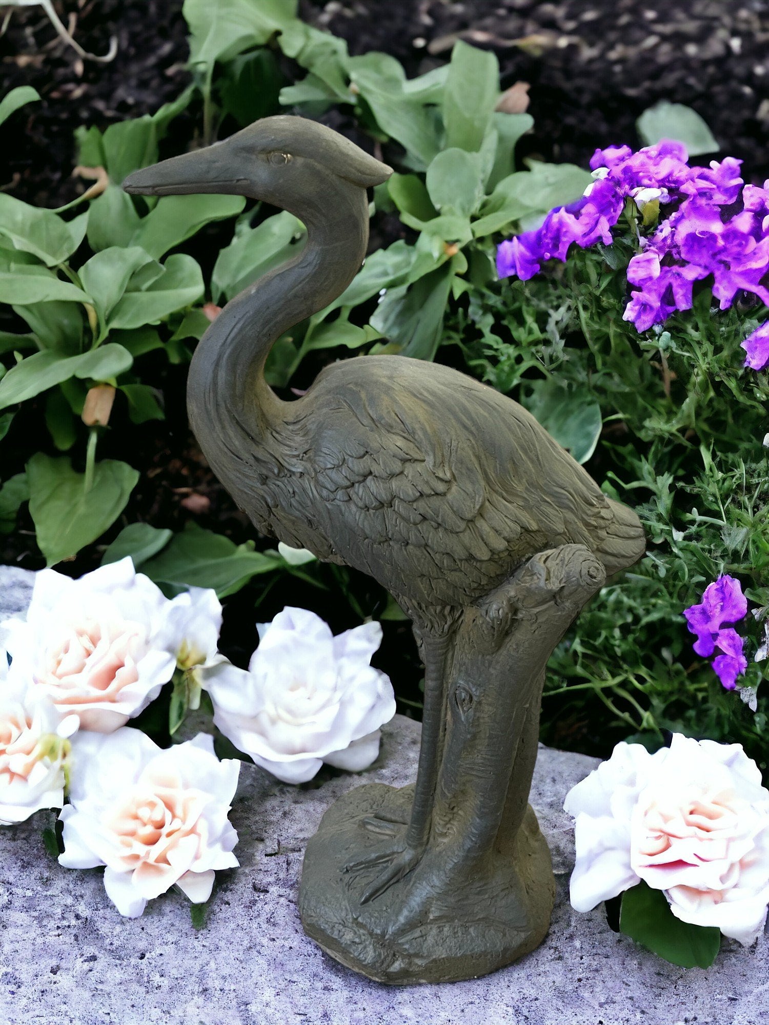 The Stone Effect Grey Heron Statue stands majestically on a pedestal, encircled by blooming white roses and violet flowers, all set against a lush green backdrop. The heron's intricately detailed feathers and elegant posture craft a tranquil outdoor scene, making it an ideal piece for garden decor.