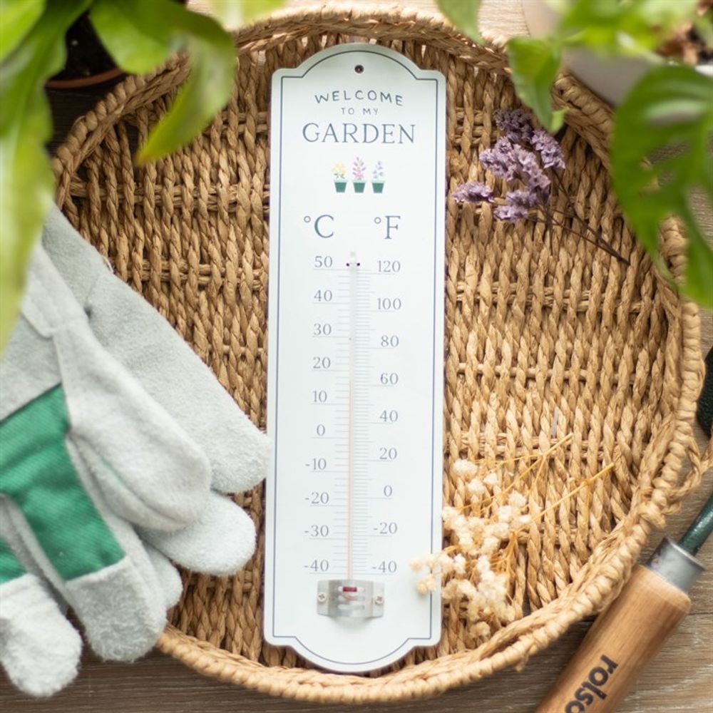 The "Welcome to My Garden Wall Thermometer" makes an ideal gift for gardeners, showcasing both Celsius and Fahrenheit on a woven mat. Encircled by gardening gloves, a small spade, and dried flowers, it’s accented by potted plants' greenery for a serene display.