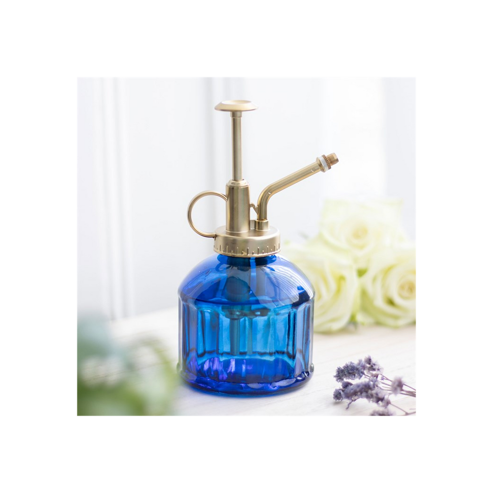 The Small Blue Glass Plant Mister, featuring a brass pump, is beautifully displayed on a light wooden surface, giving off a charming vintage vibe. In the softly blurred background, delicate lavender sprigs and white roses add an elegant touch, making it an ideal choice for any indoor garden enthusiast.