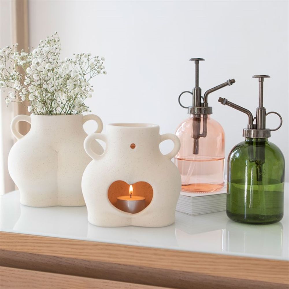 Two white, ceramic vases with abstract shapes hold a candle and flowers. Nearby, a Pink Minimalist Glass Plant Mister enhances the wooden surface, complementing the minimalist elegance of this indoor garden setup.