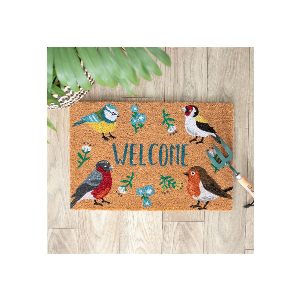 A Natural British Garden Birds Doormat features colorful birds and lush leaves on a wooden floor. To the right, a small garden fork lies nearby, and potted plant leaves elegantly extend into the top left corner of this charming scene.