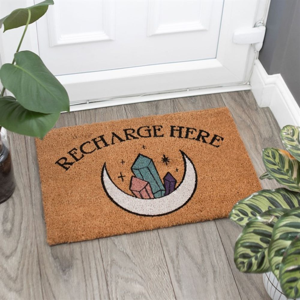 The Natural Recharge Here Crystal Doormat showcases an enchanting design with a mystical moon and vibrant crystals. It features the text "Recharge Here" and complements a white door beautifully, flanked by two lush green houseplants on a tiled floor.