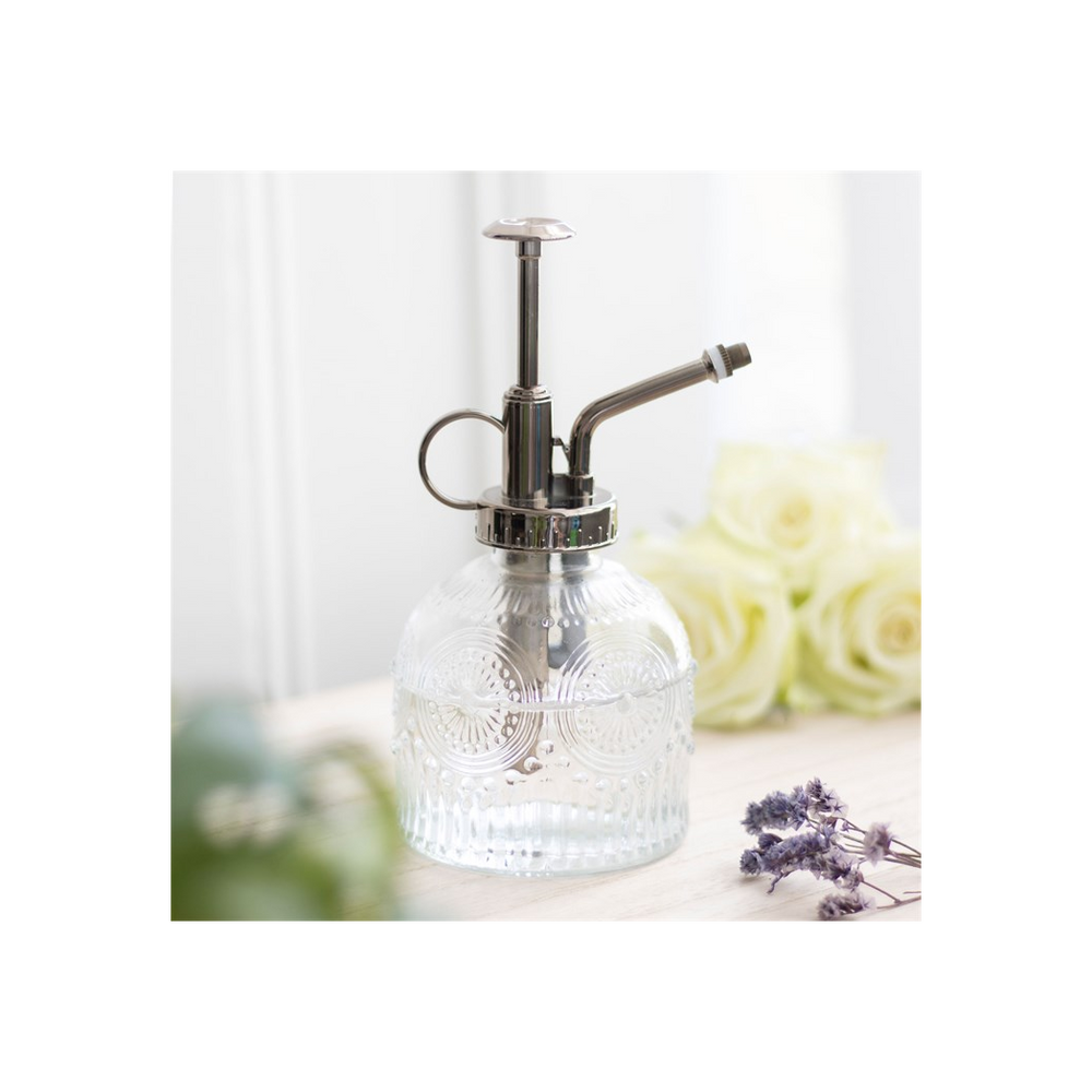 The Small Patterned Glass Plant Mister, featuring a metallic pump, sits on a light wooden surface. In the blurred background, pale yellow roses and lavender sprigs evoke the charm of an indoor garden.