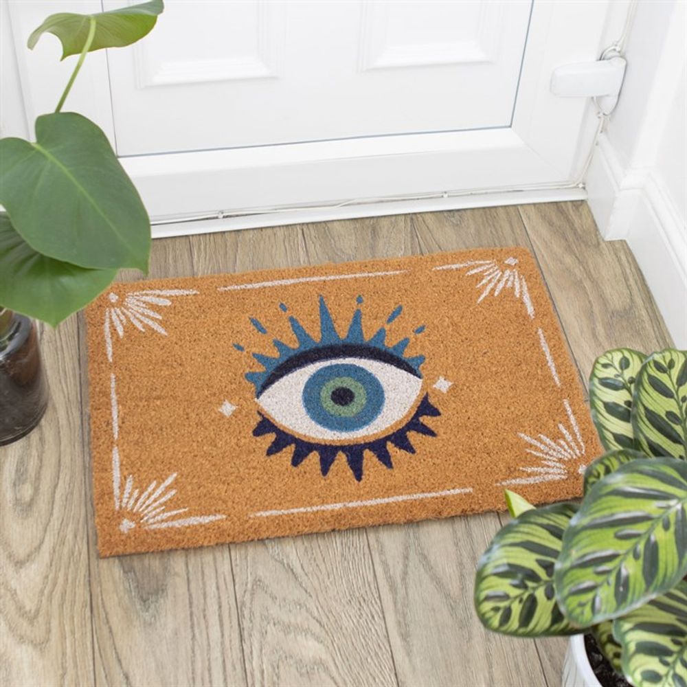 The Natural All Seeing Eye Doormat showcases a whimsical eye design with blue, green, and black tones at its center, accented decoratively in the corners. It adorns a wooden floor by a white door, accompanied by charming potted plants.