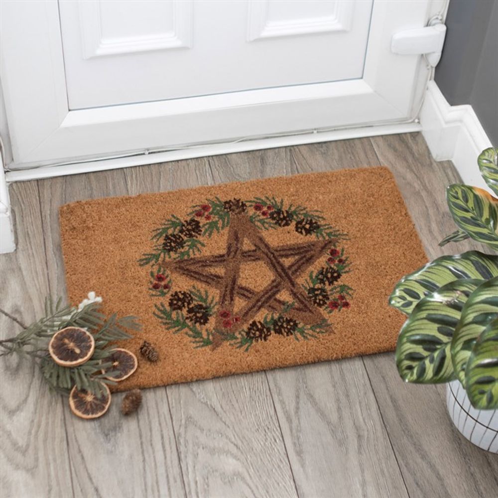 The Natural Winter Solstice Pentagram Doormat features a rustic pentagram crafted from branches at its center, accompanied by pinecones and berries. It lies on a wooden floor next to a white door, embodying the spirit of the Winter Solstice. A nearby potted plant and decorative bundle of dried fruits enhance the festive ambiance.