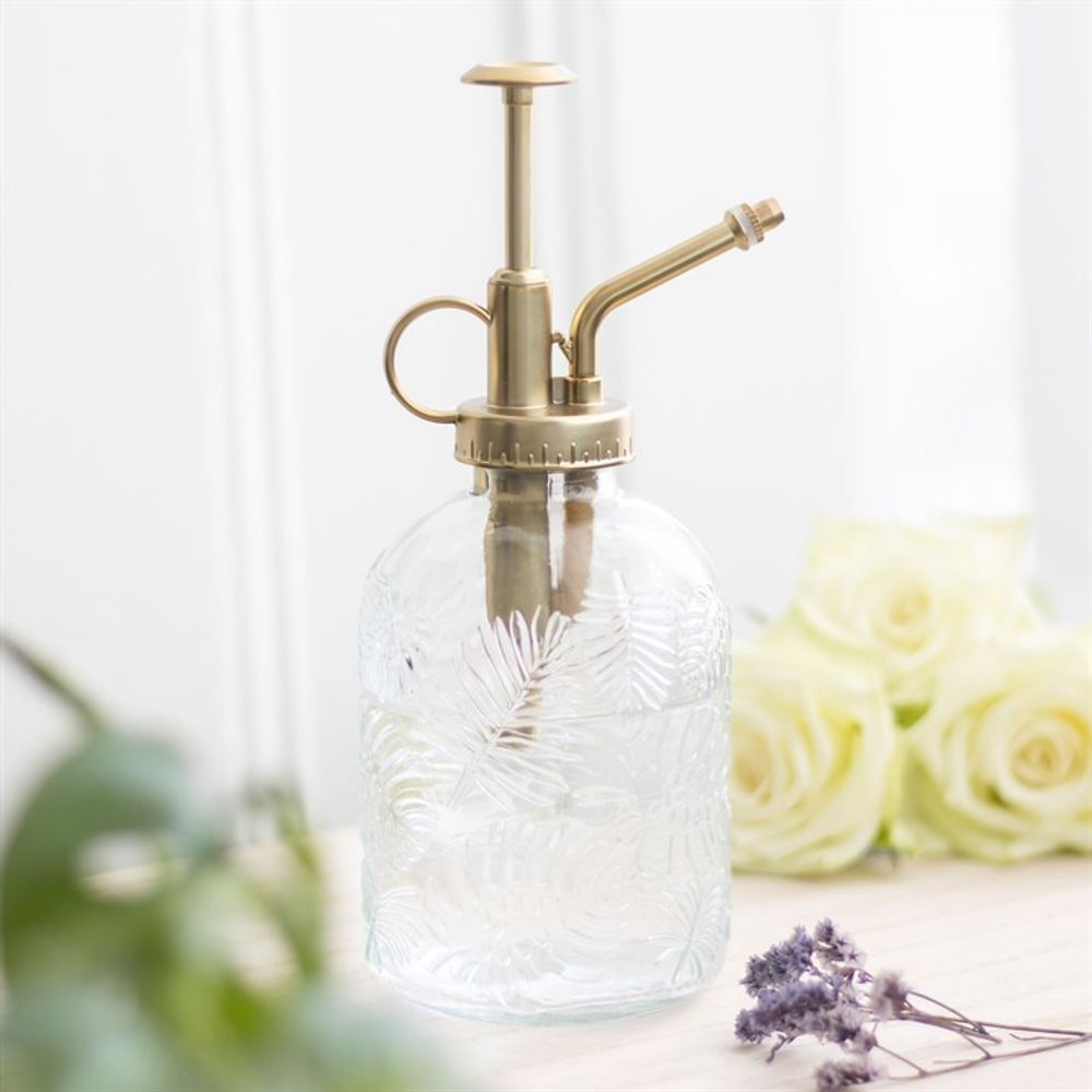The Large Leaf Print Glass Plant Mister, adorned with textured glass and a brass-colored pump, rests on a light surface. In the blurry background, white roses and sprigs of lavender enhance the charm of the indoor garden setting.