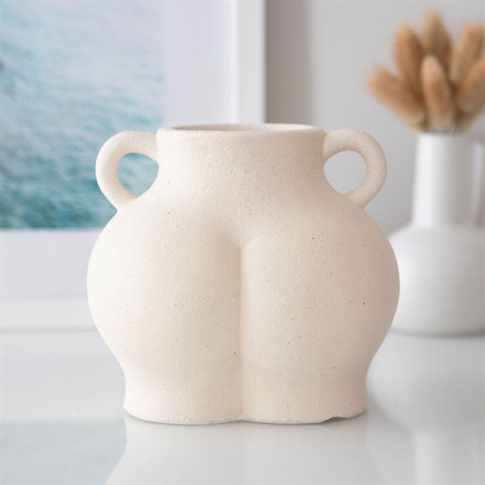 The Cream Speckle Bum Plant Pot features a quirky, beige ceramic design with a unique rounded shape resembling a human form. It includes two small handles and sits stylishly on a white surface, complementing the blurred background of framed art and dried plants, making it an ideal addition to any eclectic home decor.