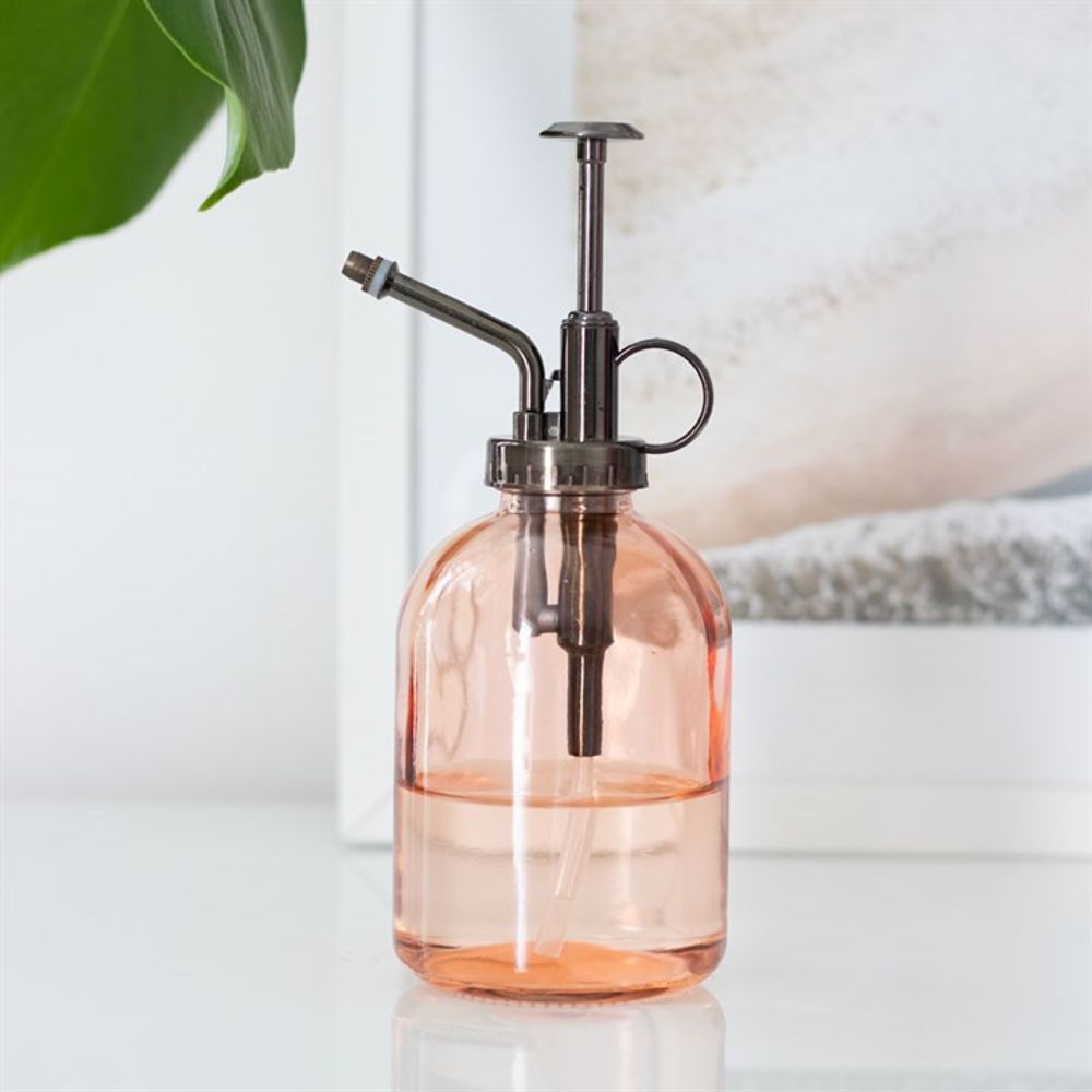 The Pink Minimalist Glass Plant Mister, featuring a transparent design with a metal pump and nozzle, is elegantly displayed on a white surface. A hint of green foliage on the left adds to its appeal as an exquisite home accessory for your indoor garden, set against a softly blurred background.