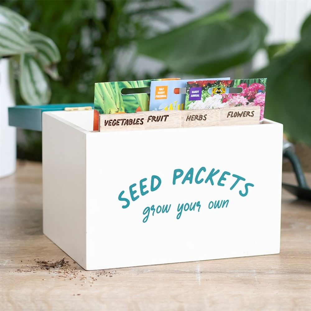 A white Seed Packet Storage Box emblazoned with "SEED PACKETS grow your own" is filled with colorful seed packets, neatly organized using labeled dividers for vegetables, fruit, herbs, and flowers. The box rests on a wooden surface scattered with soil and gardening tools, surrounded by lush green plants.