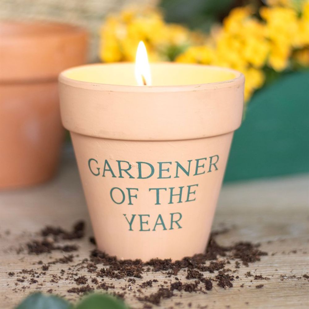 The "Gardener of the Year Citronella Candle" flickers warmly in a terracotta pot on a soil-covered wooden surface, with the soft blur of yellow flowers in the background.