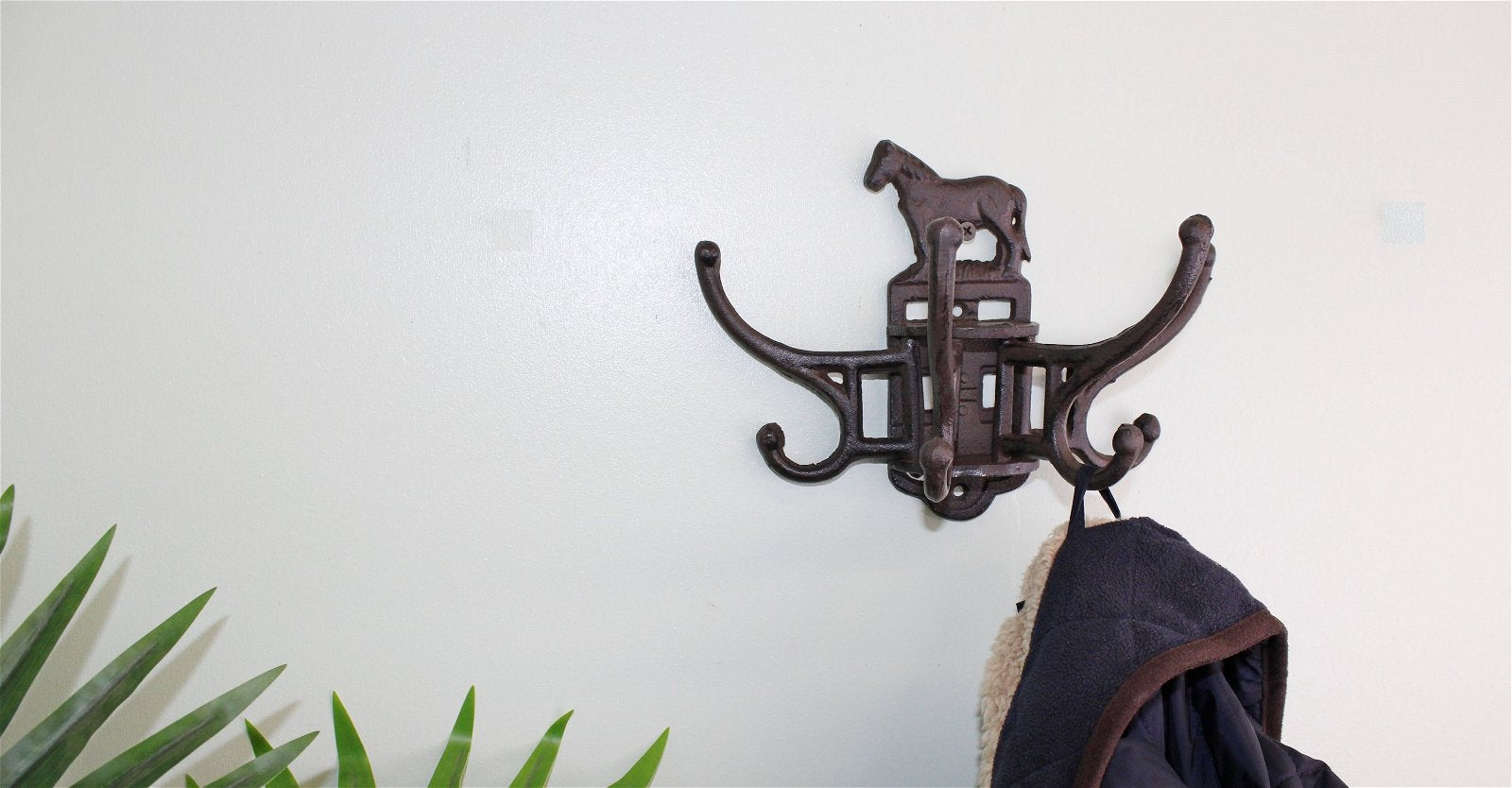 A coat hangs on a vintage-inspired Cast Iron Wall Mounted Rotating Coat Hooks with a horse design. Green plant leaves peek from the bottom left against a light wall, enhancing the industrial charm.