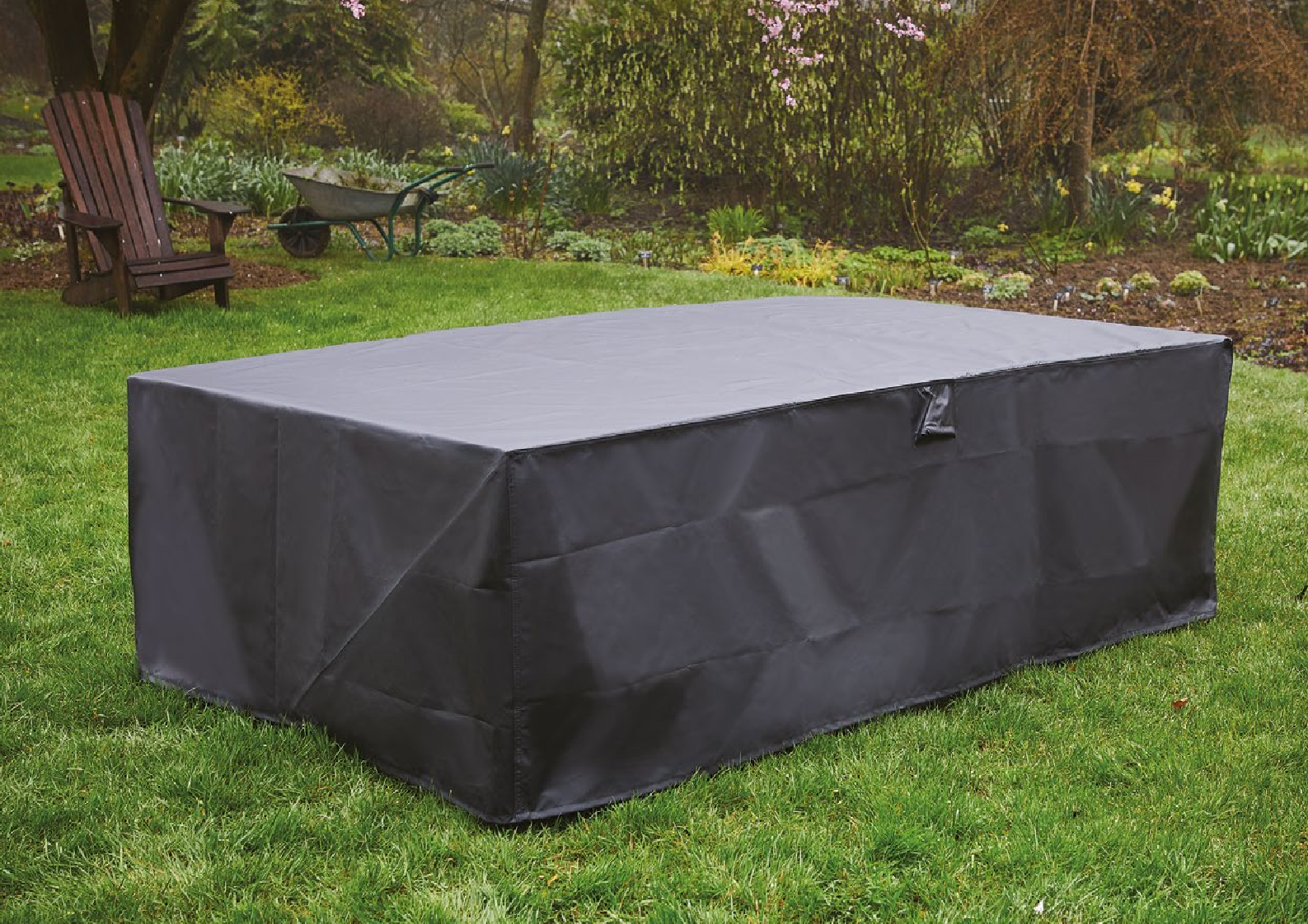 On a grassy lawn stands a large table covered with the Premium Rectangular 6-seater table cover (170 X 94 X 71 cm), surrounded by lush greenery, blooming flowers, a wooden chair, and a wheelbarrow in the background.