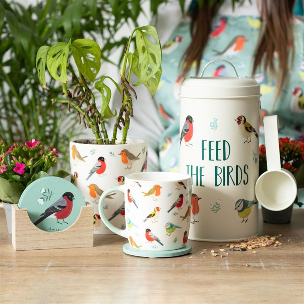 A decorative bird setup features a "Feed the Birds" tin with scoop, bird-design mugs, and blooms in a British Garden Birds Ceramic Plant Pot on a wooden table. Bird seed is scattered, garden birds add charm, and someone in a bird-patterned shirt lingers nearby.