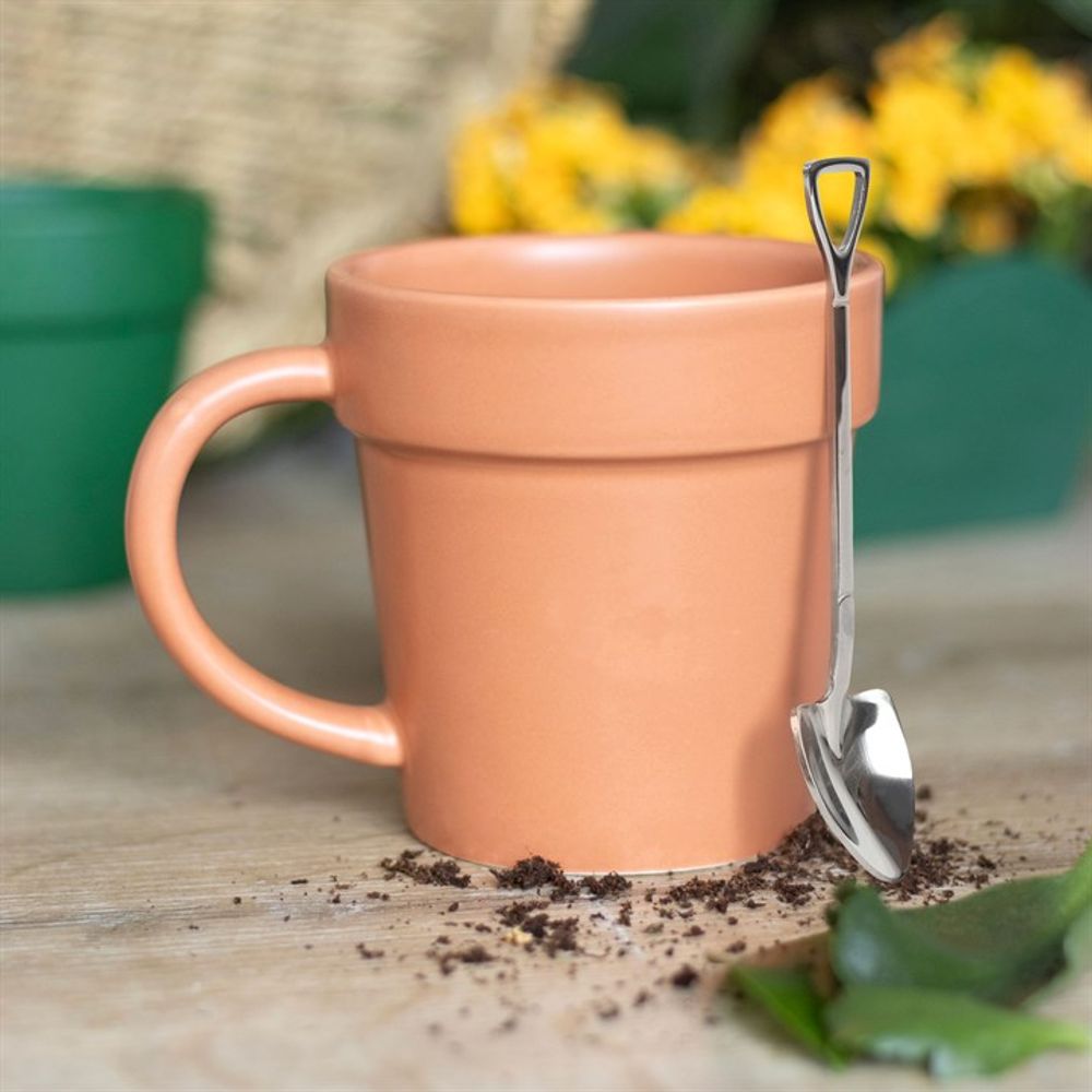 A Plain Plant Pot Ceramic Mug, resembling a terracotta pot, rests on a wooden surface with its accompanying mini shovel spoon, designed like a garden spade, leaning against it. Nearby, scattered potting soil and a green leaf add to the scene. In the background, yellow flowers and a green pot subtly capture the essence of a garden tea break.