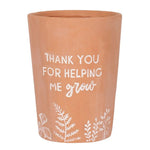 The Thank You For Helping Me Grow Terracotta Plant Pot features white text saying "Thank you for helping me grow" and charming white leaf and plant illustrations along the bottom.