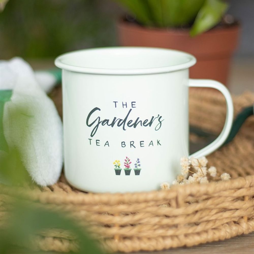A Gardener's Tea Break Enamel Mug, adorned with charming botanical illustrations, rests on a woven mat. In the blurred background, a potted plant and gardening glove hint at the scene of a keen gardener’s peaceful pause.