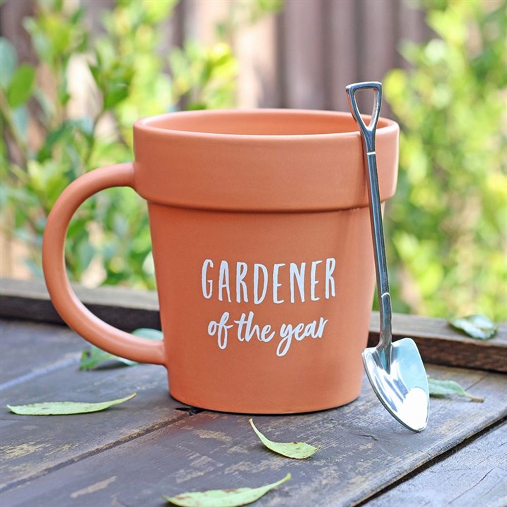The Gardener of the Year Pot Mug, shaped like a terracotta pot, sits on a wooden table with its accompanying shovel-shaped spoon resting on the rim. Scattered green leaves and blurred foliage create a charming backdrop.