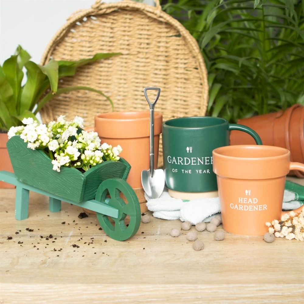 A charming garden display includes a Green Resin Wheelbarrow Planter with white flowers, a small shovel, mugs labeled "Gardener of the Year" and "Head Gardener" on a wooden table. Terracotta pots, lush plants, and a wicker basket enrich the background.