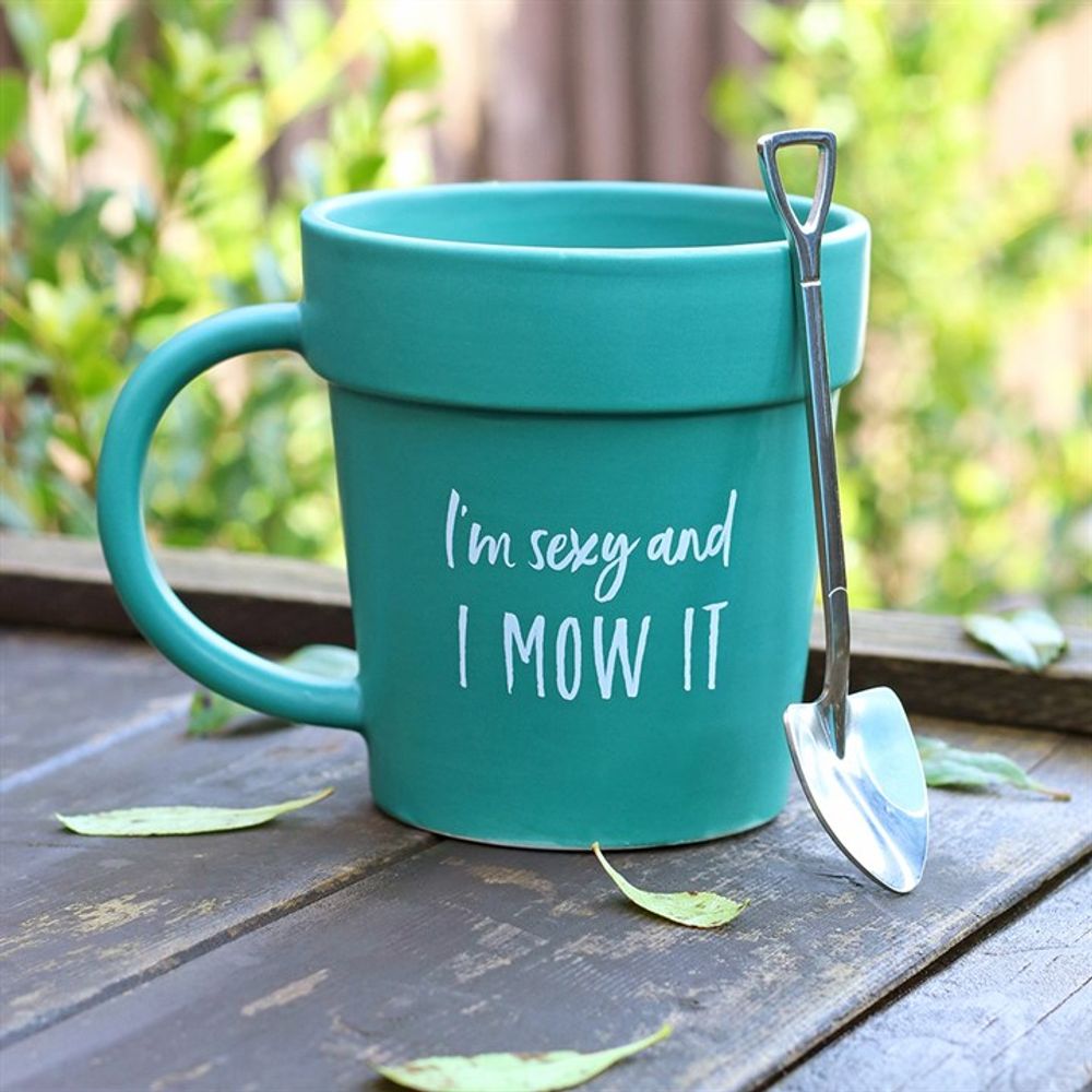 The Sexy and I Mow It Pot Mug and Shovel Spoon, a turquoise ceramic terracotta mug with a gardening theme, sits on a wooden surface surrounded by leaves. The mug showcases white text that reads, "I'm sexy and I mow it," while a small shovel-shaped teaspoon rests on its handle. Green foliage is visible in the background.