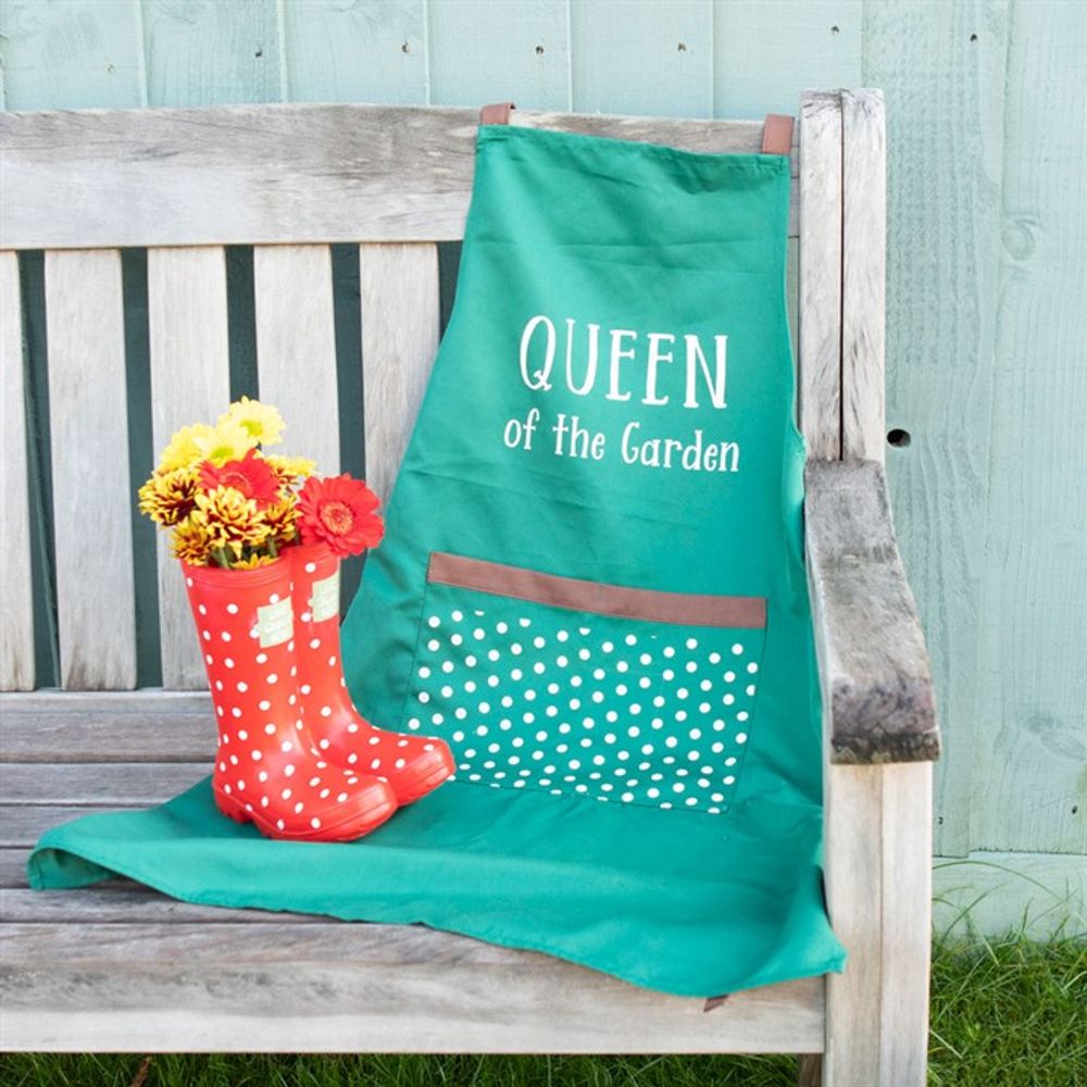 A green apron featuring the text "Queen of the Garden" hangs on a wooden bench, while a Red Welly Boot Planter displays a vibrant bouquet of yellow and red flowers. The charming turquoise wooden wall in the background completes this perfect garden decoration.