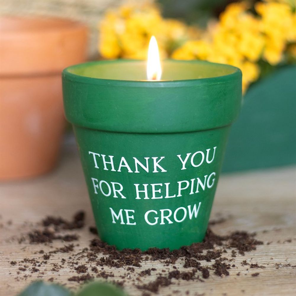 A lit "Thank You for Helping Me Grow" Citronella Candle in a stylish ceramic plant pot rests on scattered soil. Yellow flowers bloom around it, creating a welcoming outdoor ambiance.