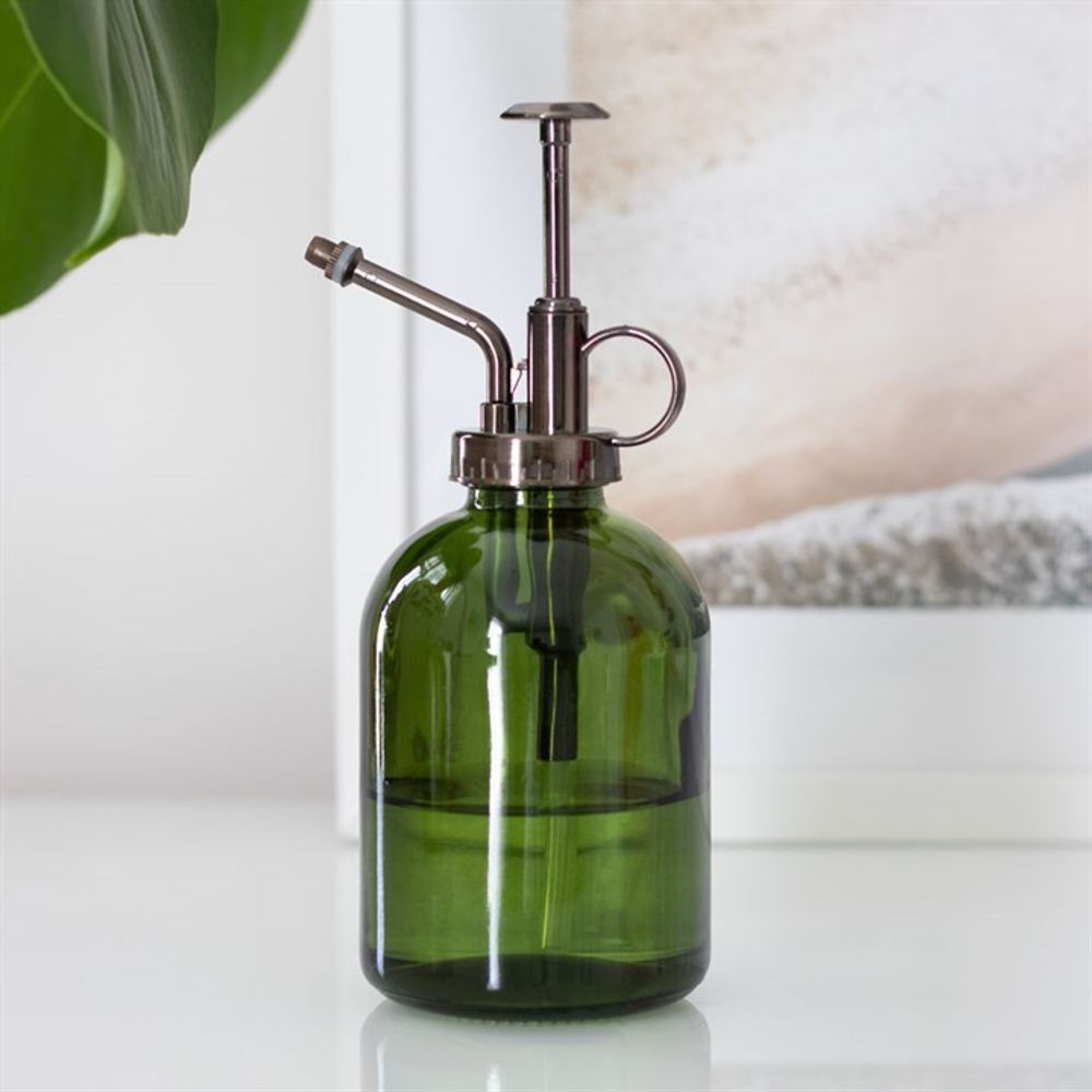 The Green Minimalist Glass Plant Mister, featuring a moss green glass body and a metal nozzle with a pump top, rests on a white surface, ideal for an indoor garden. A large green leaf is visible in the foreground, with a framed picture in the background.