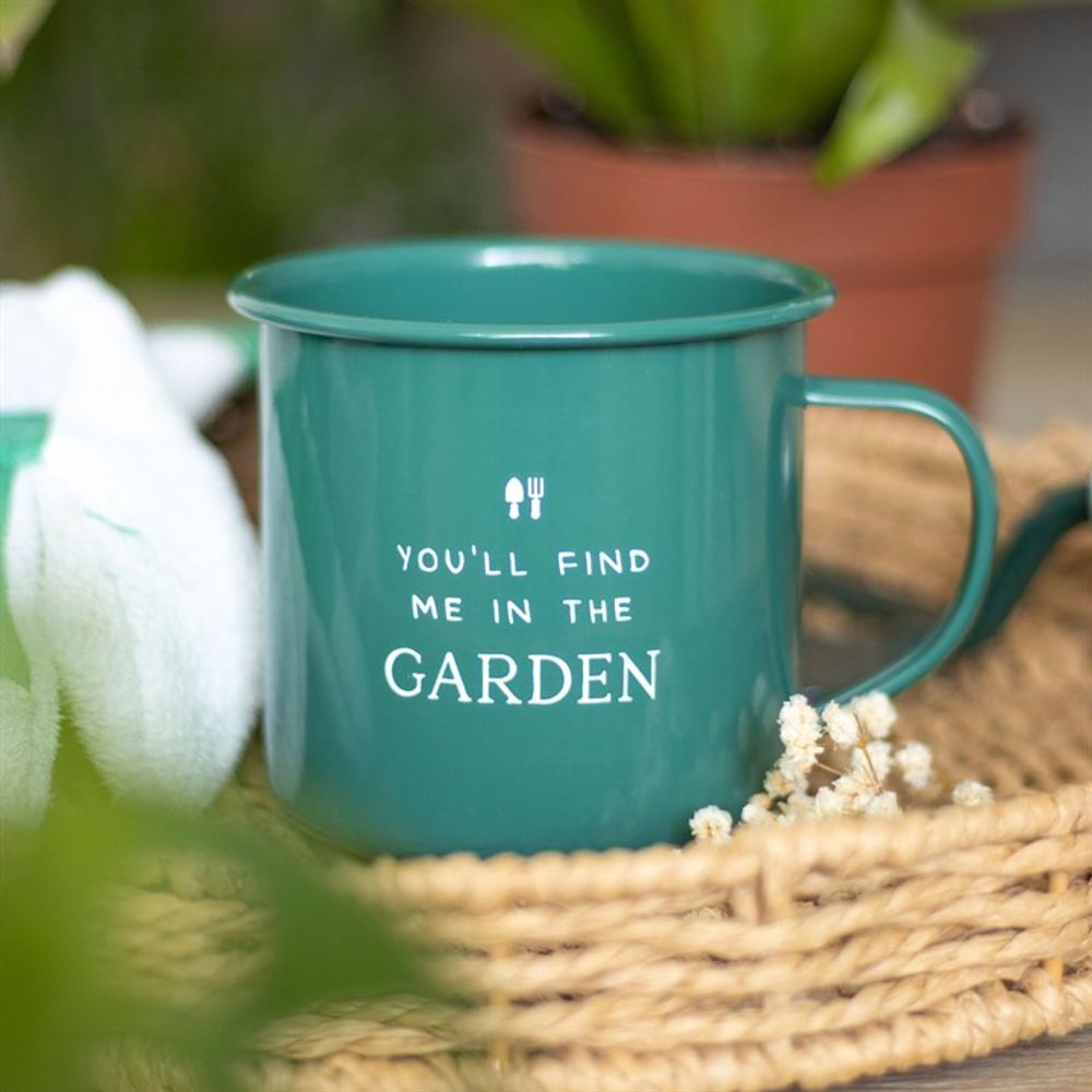 A forest green enamel mug reading "YOU'LL FIND ME IN THE GARDEN" with gardening tool icons sits in a woven basket, with a potted plant and fabric item blurred in the background, making it an ideal garden accessory. Product: Find Me in the Garden Enamel Mug.
