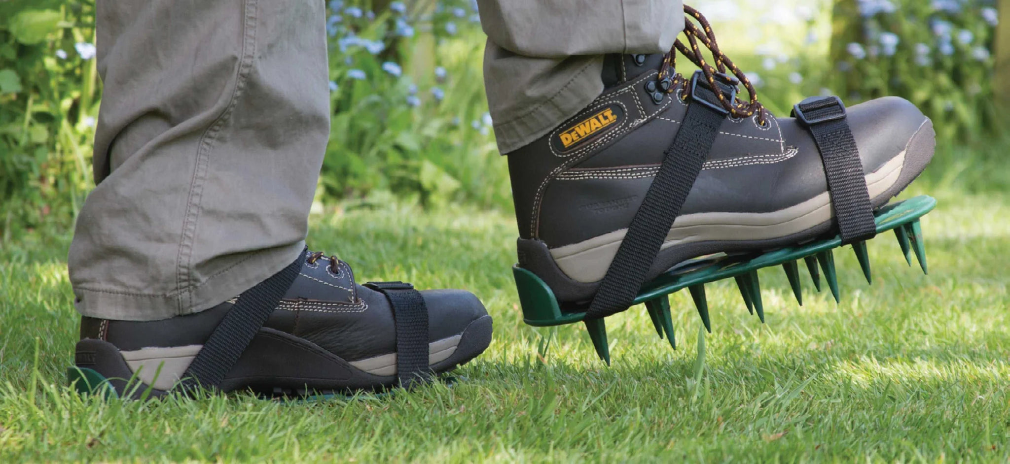 A person steps onto green grass while wearing ProSpike Lawn Aerator Shoes with Plastic Straps, securely fastened over their brown and beige boots. The lawn aerator shoes are equipped with multiple sharp cross-shaped spikes on the bottom. Greenery and blurred blue flowers can be seen in the background.