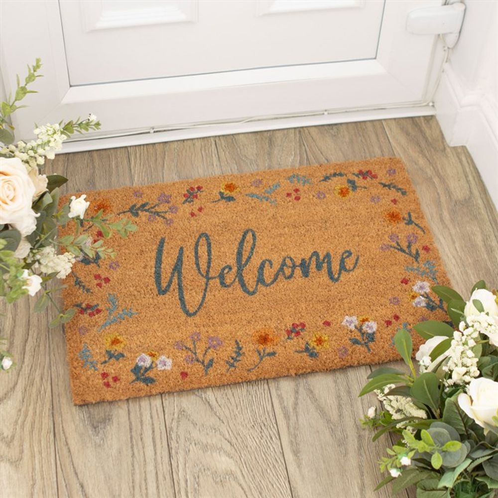 The Natural Botanical Welcome Doormat, featuring a welcome text in cursive, lies in front of a white door on a wooden floor. The beige mat is accented with colorful floral designs along the borders. Surrounding the mat are potted plants with white flowers and green leaves.