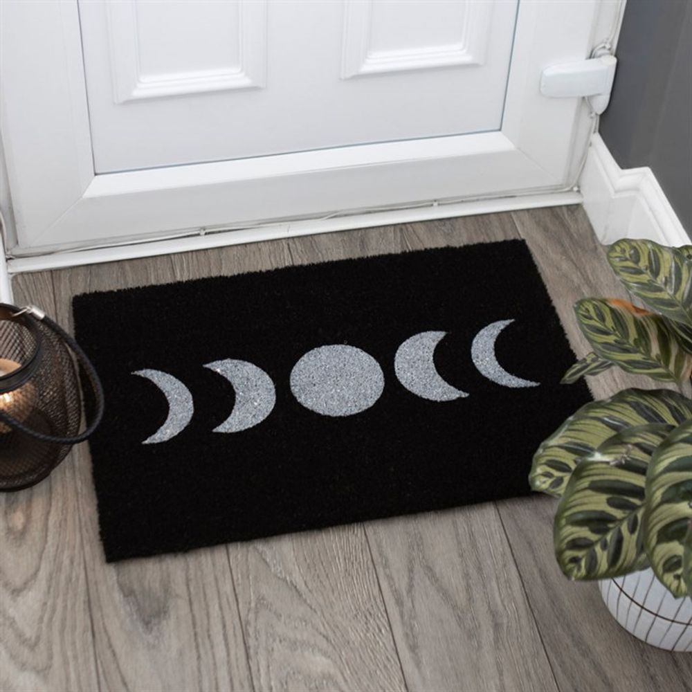 A mystical touch graces the entryway with the Black Moon Phase Doormat set before a pristine white door. Adorned with a white moon phase design on its rectangular black surface, it is flanked by a potted plant with variegated green leaves on the right and a round lantern on the left, resting on light wood flooring.