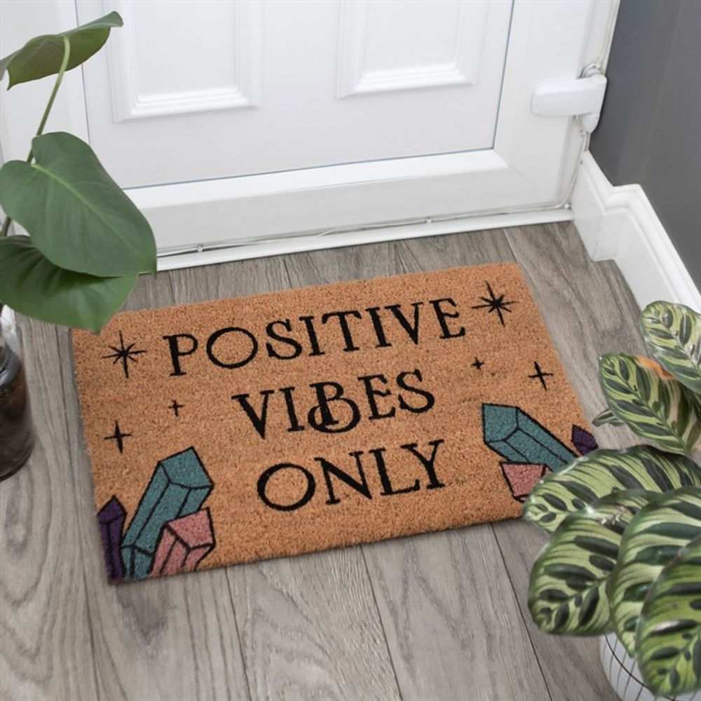 A "Natural Positive Vibes Only Doormat" placed in front of a white door displays the cheerful message in black letters. The mat is decorated with illustrations of colorful magic crystals and small star shapes, promoting love and happiness. Visible in the surroundings are parts of two green leafy plants on a gray wooden floor.