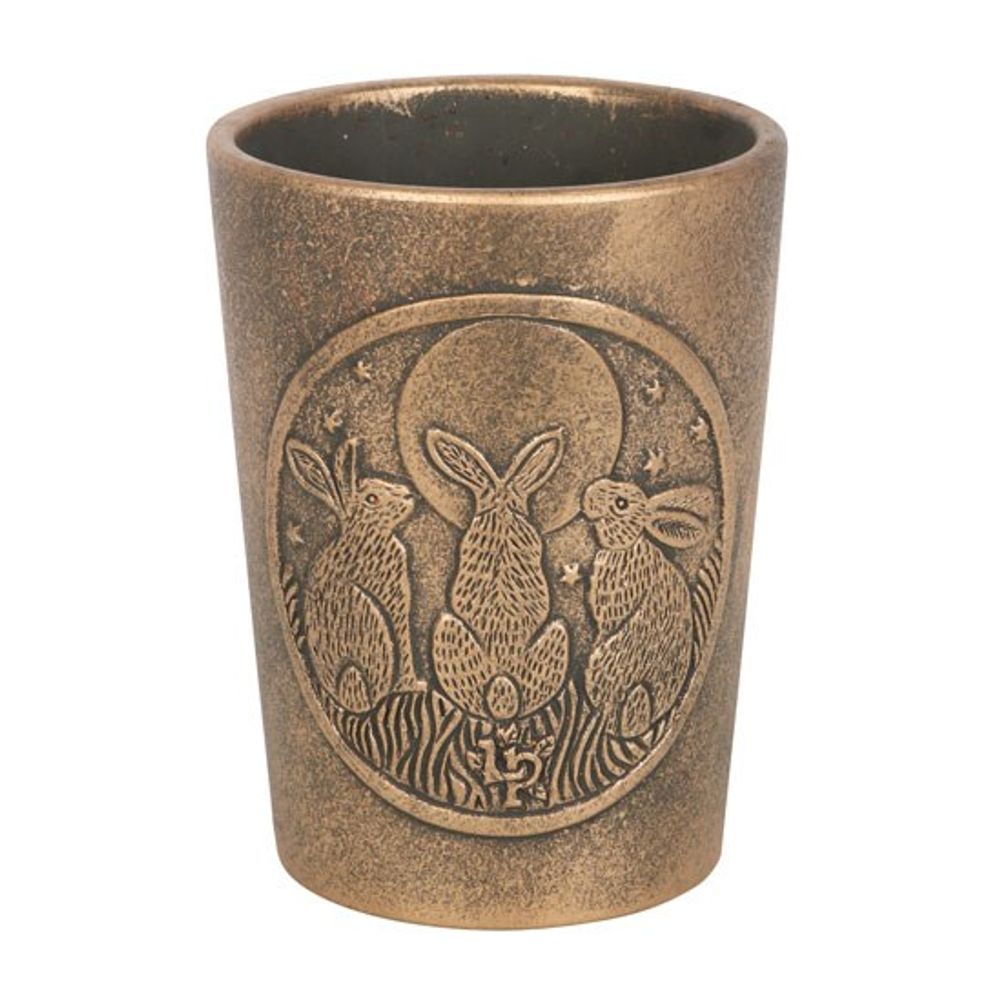The Moon Shadows Bronze Terracotta Plant Pot by Lisa Parker is a bronze-colored cylindrical piece, featuring an embossed design of three rabbits sitting under a full moon, surrounded by stars and grass. Reminiscent of Lisa Parker's Moon Shadows artwork, the intricate details give the pot an elegant and rustic appearance.