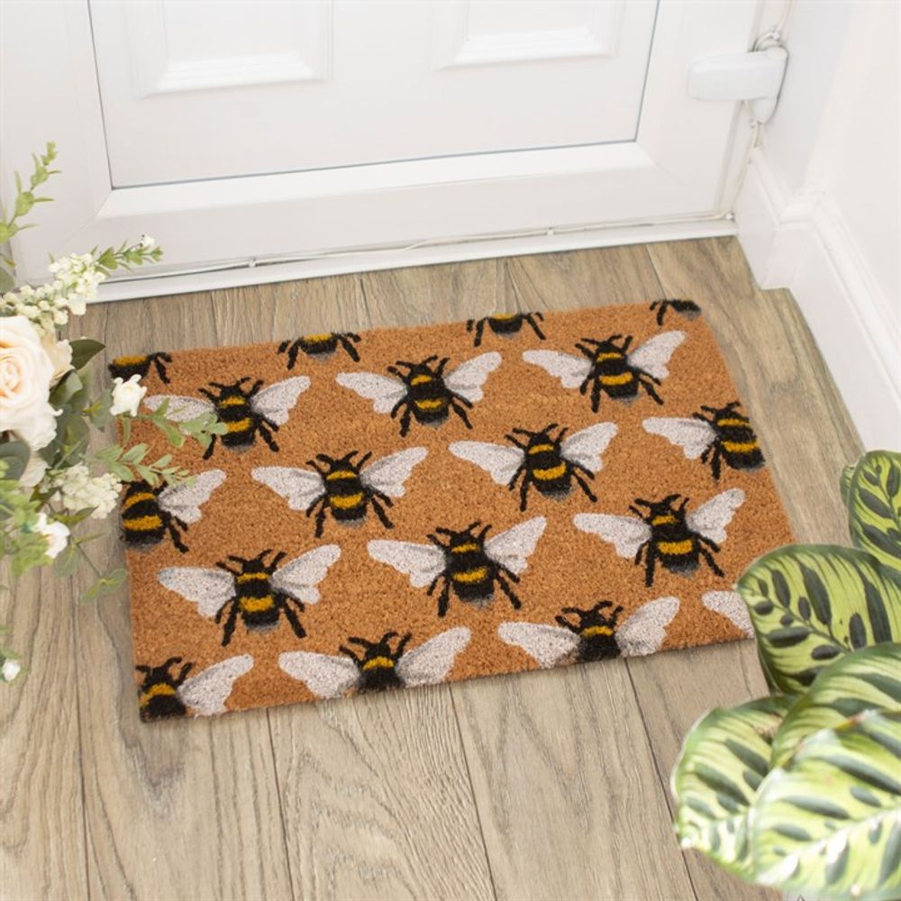 A Natural Bee Print Doormat with an all-over pattern of black and yellow bees, each featuring white wings, is placed in front of a white door. The doormat rests on wood flooring, surrounded by greenery including plants and flowers. Perfect for indoor use, it adds a charming touch to your home.