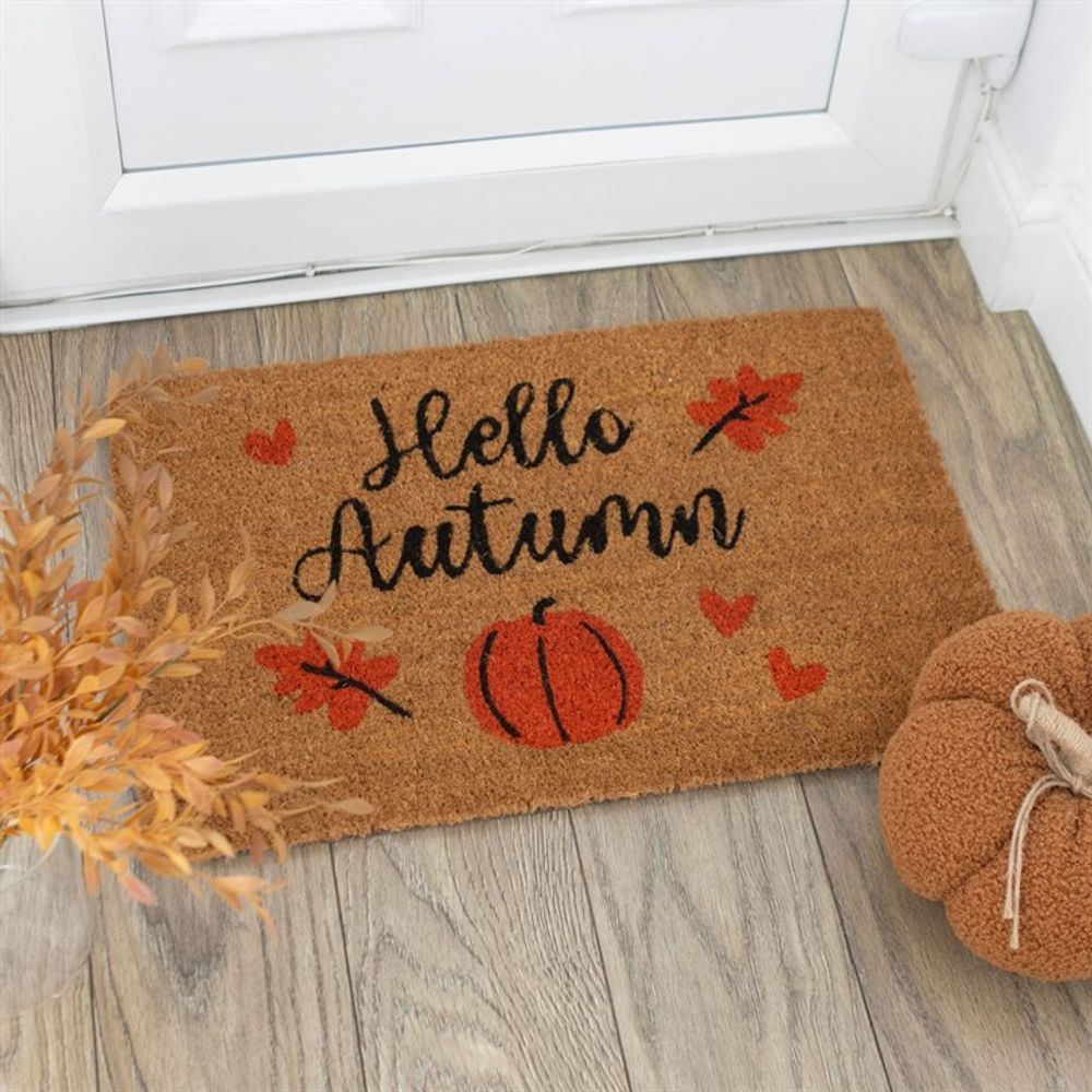 A Natural Hello Autumn Doormat with "Hello Autumn" written in black cursive is placed by a white door. The mat showcases a vibrant design of red leaves and pumpkins, adding festive charm. Nearby, dried leaves and a knitted pumpkin decoration rest on the wooden floor.