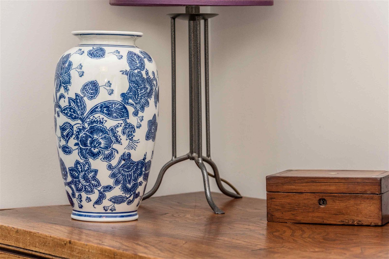 A tall floral vase shown with a lamp on a wooden table.