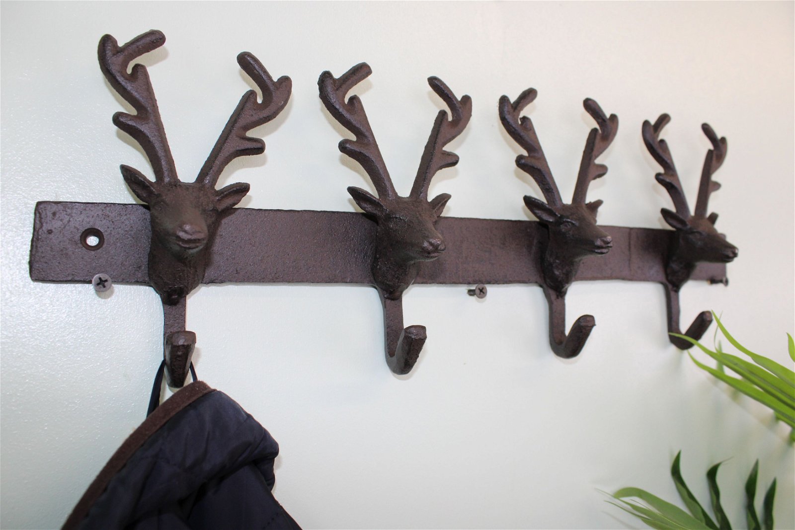 A wall-mounted wrought iron coat rack features four Rustic Cast Iron Wall Hooks shaped like reindeer heads, with one hook holding a dark jacket. Positioned beside this industrial-style decor is a green plant, adding a touch of nature to your space storage accessories.