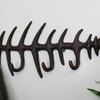 The Rustic Cast Iron Wall Hooks, Fish Skeleton, serve as both functional and decorative wall-mounted coat hooks. Featuring a design with the head, ribs, and tail of a fish skeleton, this hook includes several pegs ideal for small space storage accessories. A dark-colored coat hangs on one of the hooks near a partially visible green plant.