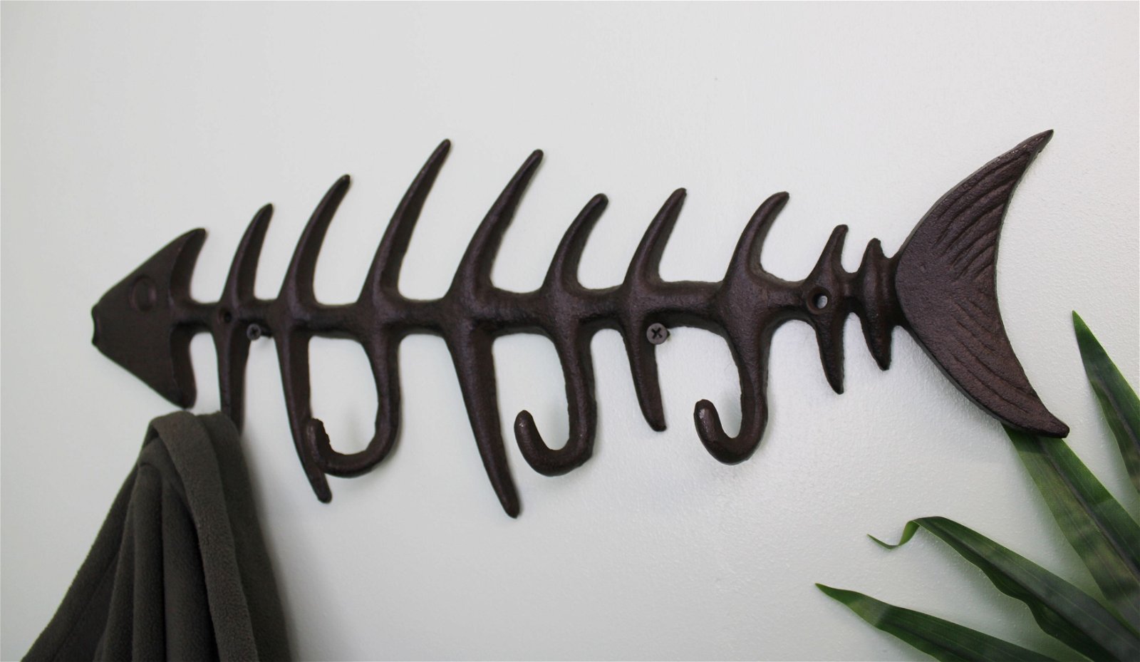 The Rustic Cast Iron Wall Hooks, Fish Skeleton, serve as both functional and decorative wall-mounted coat hooks. Featuring a design with the head, ribs, and tail of a fish skeleton, this hook includes several pegs ideal for small space storage accessories. A dark-colored coat hangs on one of the hooks near a partially visible green plant.