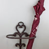 A maroon umbrella with a light brown handle is neatly stored in a Traditional Rustic Cast Iron Umbrella Stand featuring a heart-shaped design, placed against a light-colored wall near a wooden floor, ensuring a drip-free hallway.